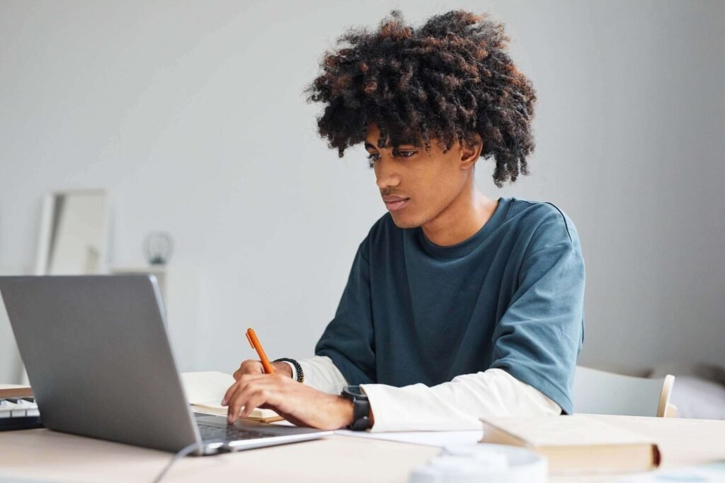 Joven con cabello rizado escribiendo en una libreta mientras mira la pantalla de su laptop en un escritorio.